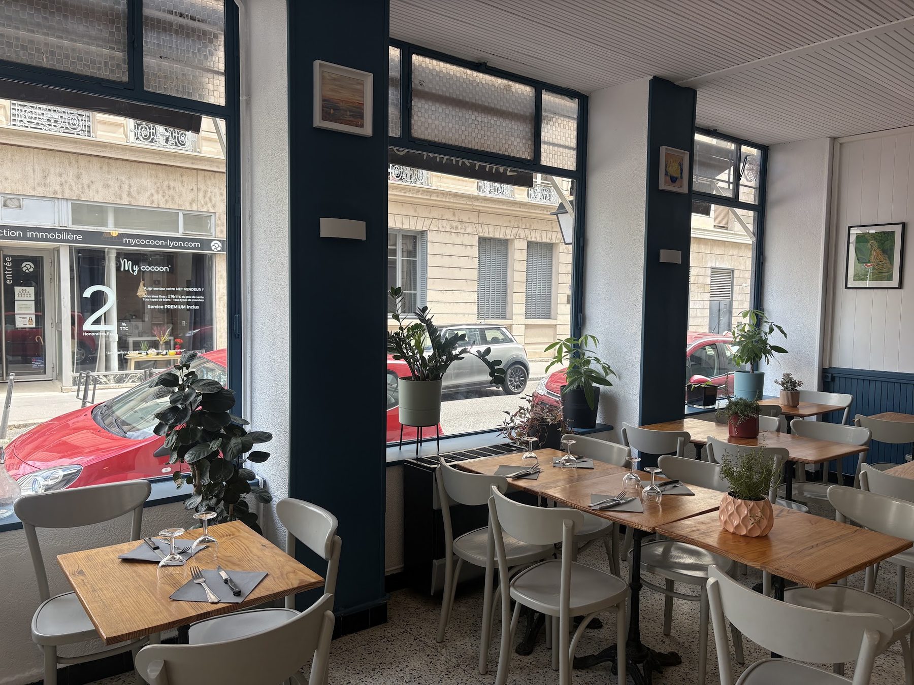 Der Gastraum des Café de la Mairie: Holztische, große marineblaue Fenster, Pflanzen